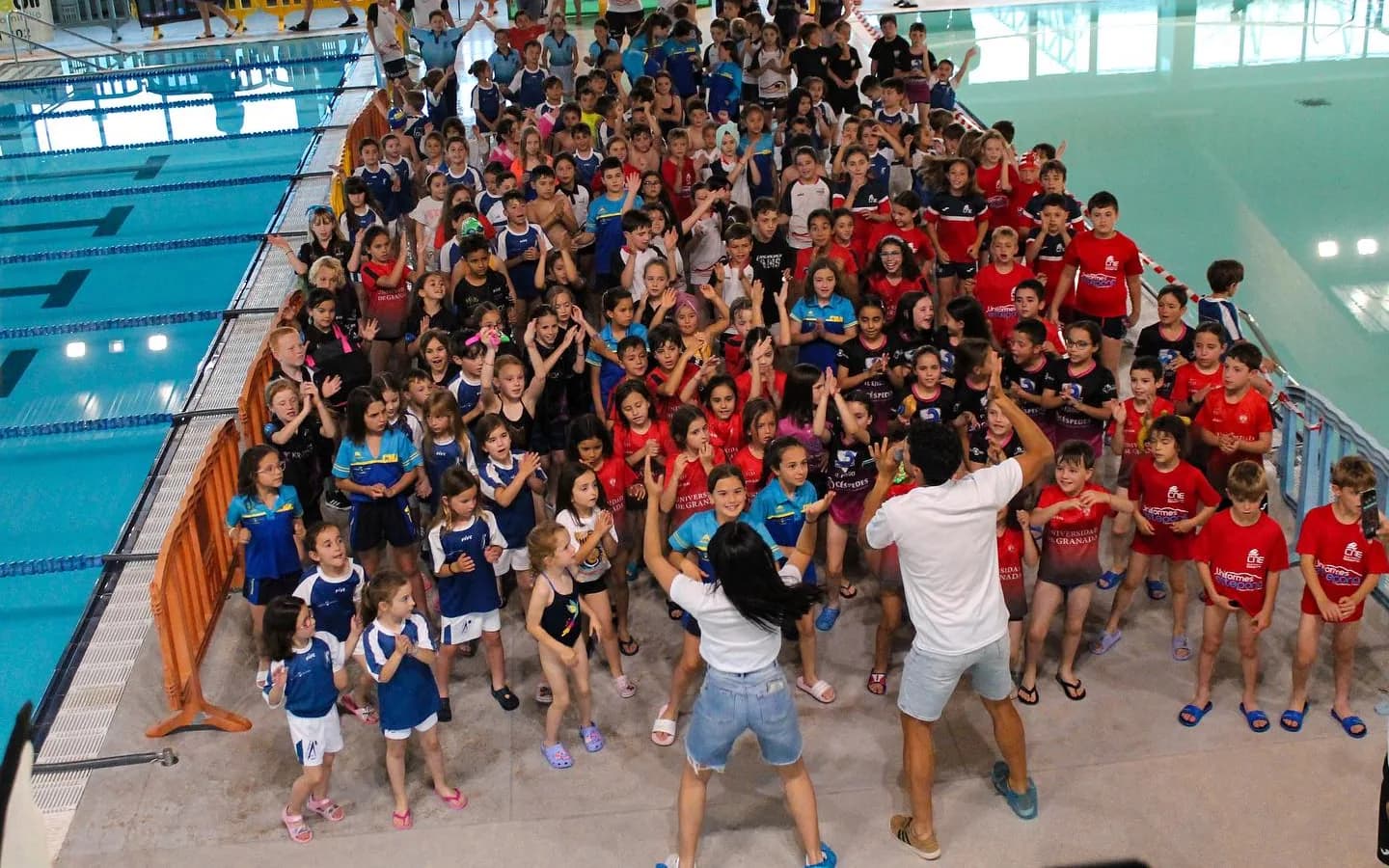 Vista desde arriba de unos doscientos niños y niñas bailando en el bordillo de la piscina de Torre del Mar con dos animadores, en el XII Open Chupete de mayo de 2024