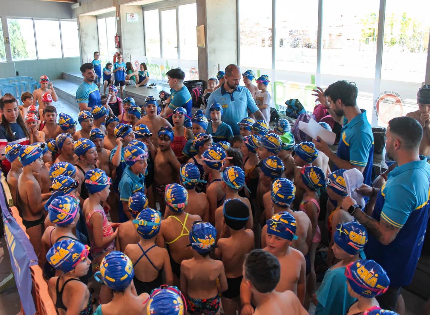 Entrenadores con muchos niños y niñas con gorros del CNA, reunidos junto a la piscina cubierta en el XII Open Chupete